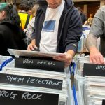 A group of people browse through vinyl records at a busy record fair. The image shows several plastic crates labeled with handwritten black signs indicating music genres and categories, including "IRON MAIDEN," "SYNTH / ELECTRONIC," "PROG ROCK," "MISC Q–T," "PUNK," and "GRUNGE / ALT ROCK." A man in the center wearing a white "Blue Flowers" t-shirt and navy hoodie is examining a record. To his right, an older man in an AC/DC t-shirt and with a tattooed arm is also browsing. The setting appears lively and crowded with music enthusiasts.