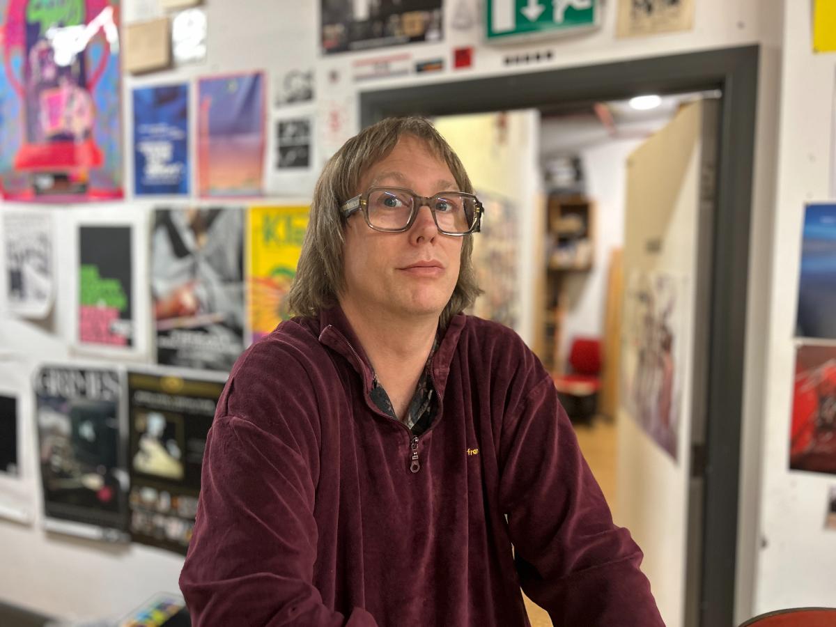 Matt Bradshaw - a person with shoulder-length hair and large glasses is standing indoors, leaning slightly forward with arms resting on a surface. They are wearing a maroon zip-up fleece. Behind them is a wall covered with various colorful posters and flyers, suggesting a creative or academic environment. An open doorway reveals a room with shelves and a red chair.