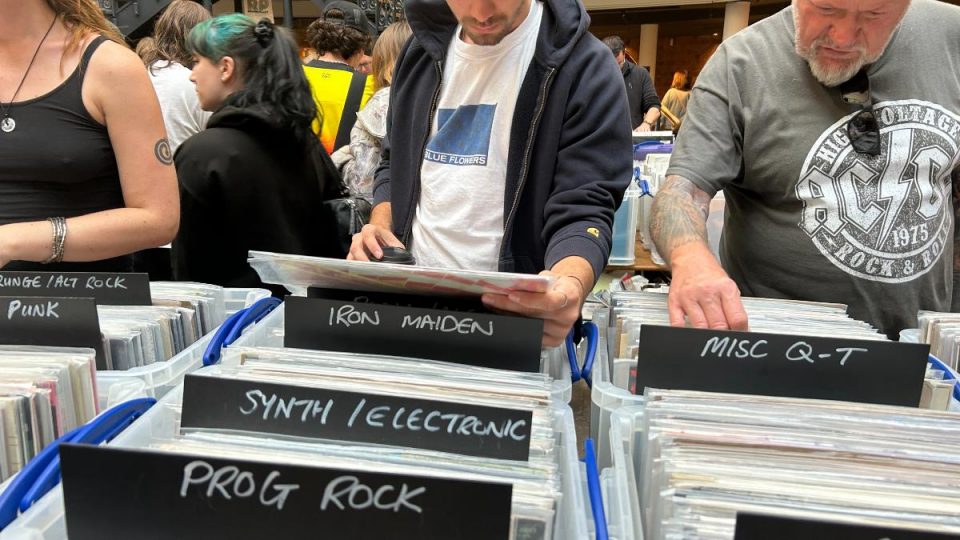 A group of people browse through vinyl records at a busy record fair. The image shows several plastic crates labeled with handwritten black signs indicating music genres and categories, including "IRON MAIDEN," "SYNTH / ELECTRONIC," "PROG ROCK," "MISC Q–T," "PUNK," and "GRUNGE / ALT ROCK." A man in the center wearing a white "Blue Flowers" t-shirt and navy hoodie is examining a record. To his right, an older man in an AC/DC t-shirt and with a tattooed arm is also browsing. The setting appears lively and crowded with music enthusiasts.