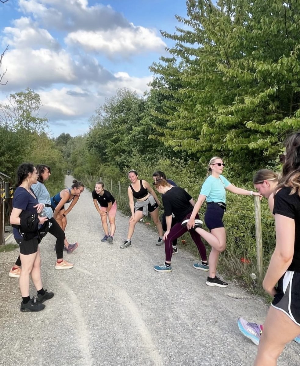 The Strega Women's Running Club in Leeds warming up by a lake and trees, around thirteen of them, all in bright, athletic clothing