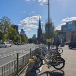 The image shows a rack of E-bikes and bikes on Woodhouse Road, Leeds with a view towards the city centre. You can see a church and high rise buildings in the background