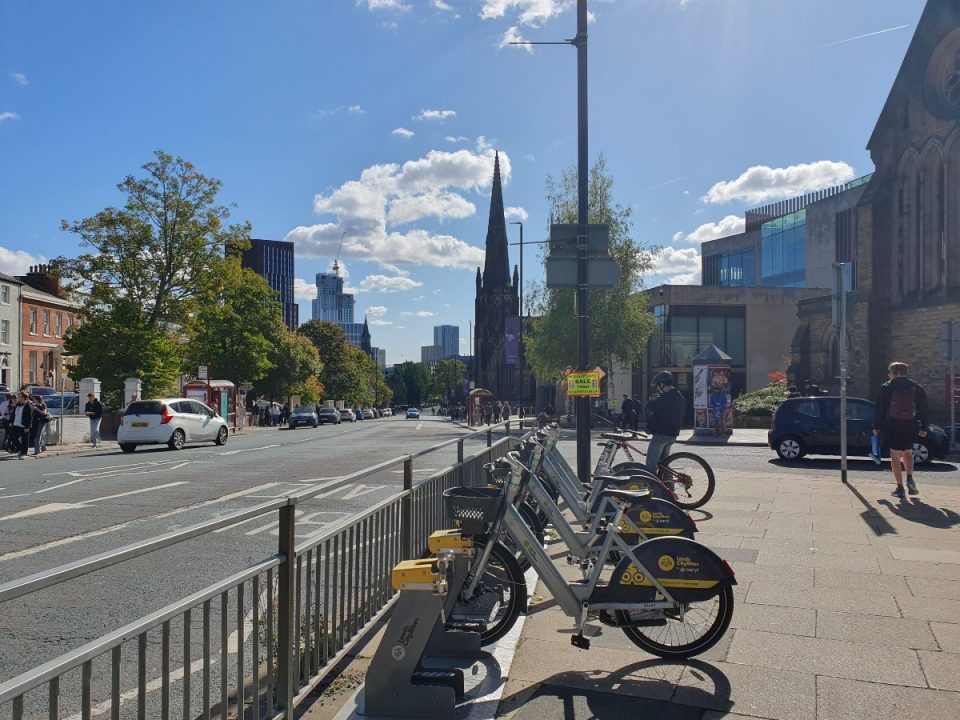The image shows a rack of E-bikes and bikes on Woodhouse Road, Leeds with a view towards the city centre. You can see a church and high rise buildings in the background