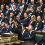 Kemi Badenoch speaking at a podium in a formal parliamentary setting, surrounded by seated MP's in suits and formal attire, with tiered benches and ornate woodwork visible in the background