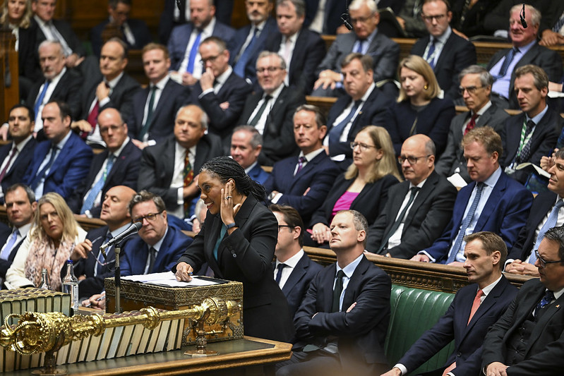 Kemi Badenoch speaking at a podium in a formal parliamentary setting, surrounded by seated MP's in suits and formal attire, with tiered benches and ornate woodwork visible in the background