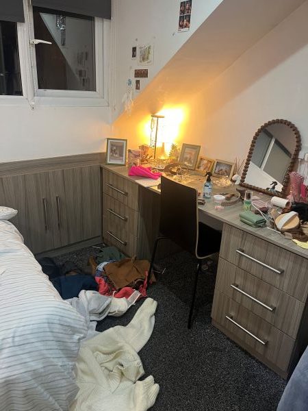 Bedroom with a bed, cluttered desk under a window, and drawers; pictures hang on the wall.