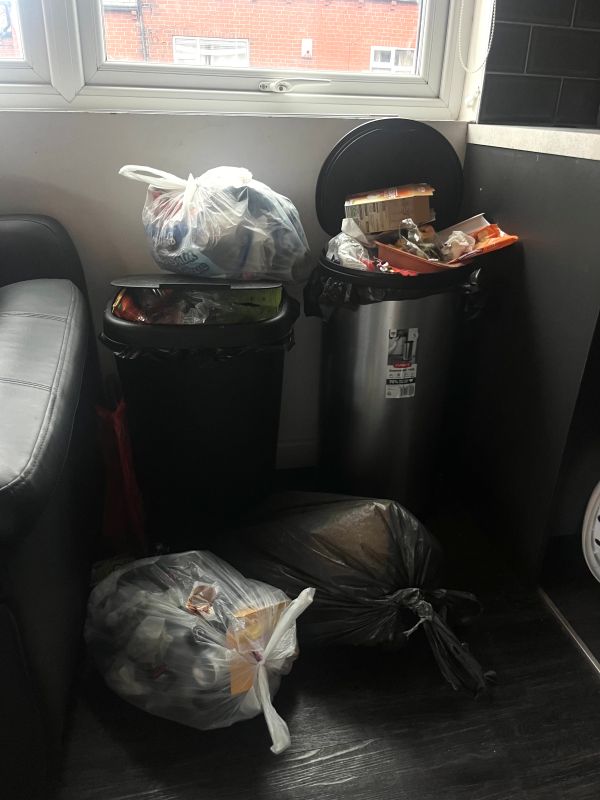  Two overflowing trash bins with extra garbage bags on the floor beside them in a room corner near a window.
