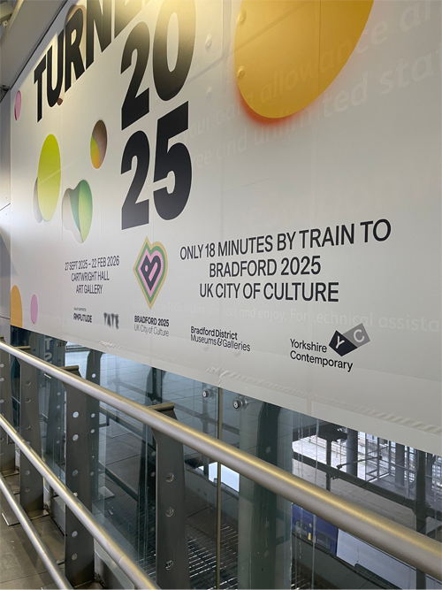Large wall advertisement inside a station promoting the Turner 2025 exhibition and Bradford 2025 UK City of Culture, stating ‘Only 18 minutes by train to Bradford 2025 UK City of Culture,’ with logos for Tate, Bradford District Museums & Galleries, and Yorkshire Contemporary.