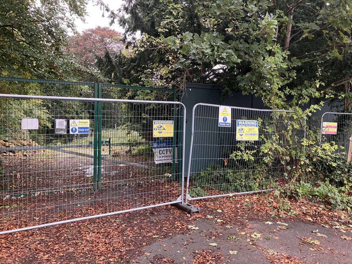 A fenced-off entrance to a wooded area with multiple warning signs including 'Danger – Keep Out,' 'CCTV in operation,' and '24/7 monitored site.' Temporary metal fencing is placed in front of a green gate, surrounded by trees and fallen autumn leaves.