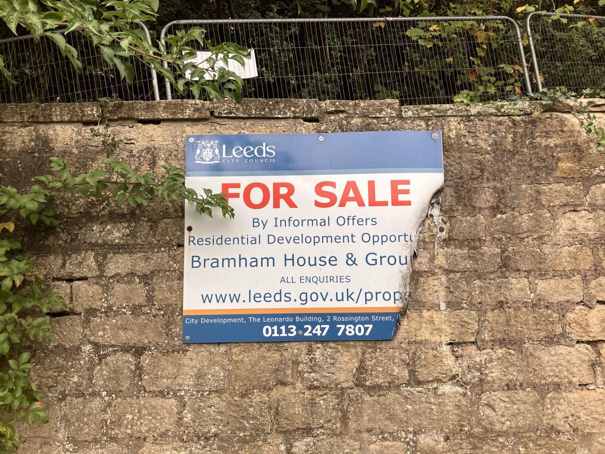 A weathered "For Sale" sign from Leeds City Council is attached to a stone wall, partially peeling away at the corner. The sign advertises a residential development opportunity for Bramham House & Grounds. It includes a website (www.leeds.gov.uk/prop ...) and contact number (0113 247 7807) for City Development at The Leonardo Building, 2 Rossington Street. There is overgrown foliage and metal fencing above the wall, with a wooded area in the background.