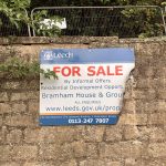 A weathered "For Sale" sign from Leeds City Council is attached to a stone wall, partially peeling away at the corner. The sign advertises a residential development opportunity for Bramham House & Grounds. It includes a website (www.leeds.gov.uk/prop ...) and contact number (0113 247 7807) for City Development at The Leonardo Building, 2 Rossington Street. There is overgrown foliage and metal fencing above the wall, with a wooded area in the background.
