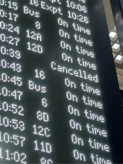 Train station platform with several passengers waiting, some with luggage, beneath a large glass and steel roof structure with multiple overhead lights and visible signage.