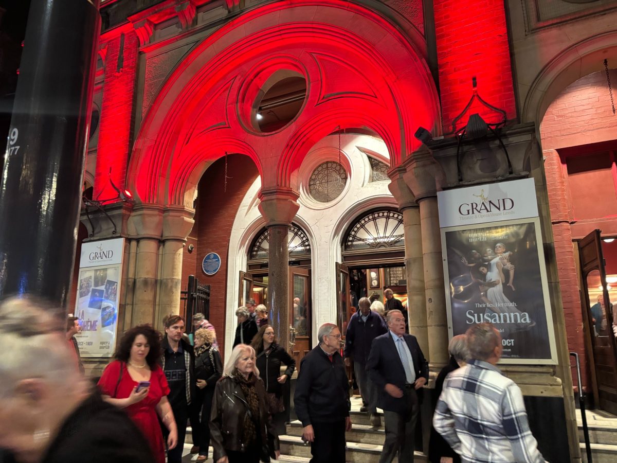 People exit a theatre under a vibrant red-lit arch. Posters on the walls announce the performance of “Susanna.”