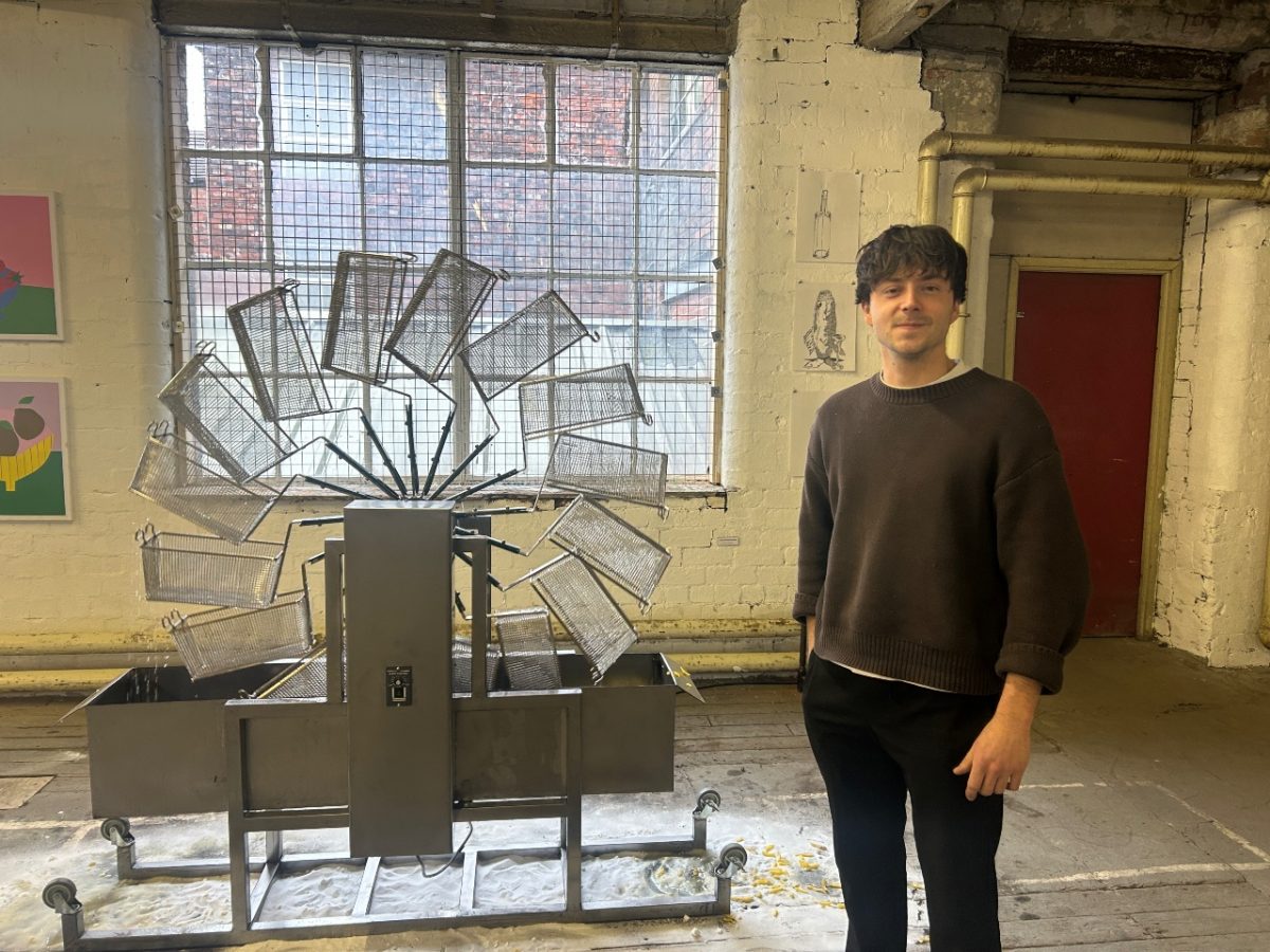 image of sculpture of a deep fat fryer mill, with baskets that turn and drop into a vat of oil. The artist, a young, white male with brown hair stands next to his piece. He is wearing black jeans and a beige jumper.