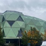 Headingley Stadium: a modern building with green geometric facades and triangular windows, surrounded by trees with autumn-coloured leaves under a cloudy sky.