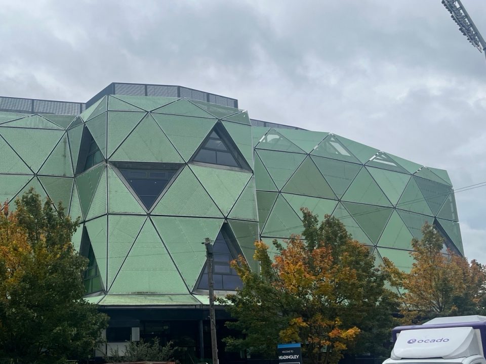 Headingley Stadium: a modern building with green geometric facades and triangular windows, surrounded by trees with autumn-coloured leaves under a cloudy sky.
