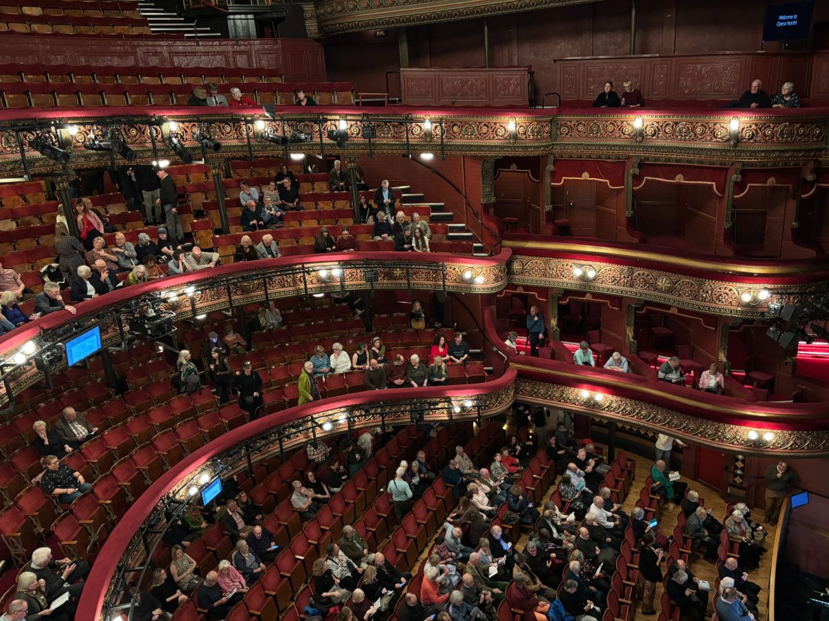 Audience members take their seats in the ornate balconies and stalls of a theater before a performance. Some seats are still empty.