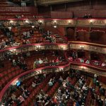 Audience members take their seats in the ornate balconies and stalls of a theater before a performance. Some seats are still empty.