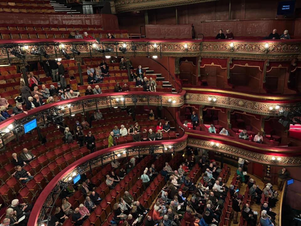 Audience members take their seats in the ornate balconies and stalls of a theater before a performance. Some seats are still empty.
