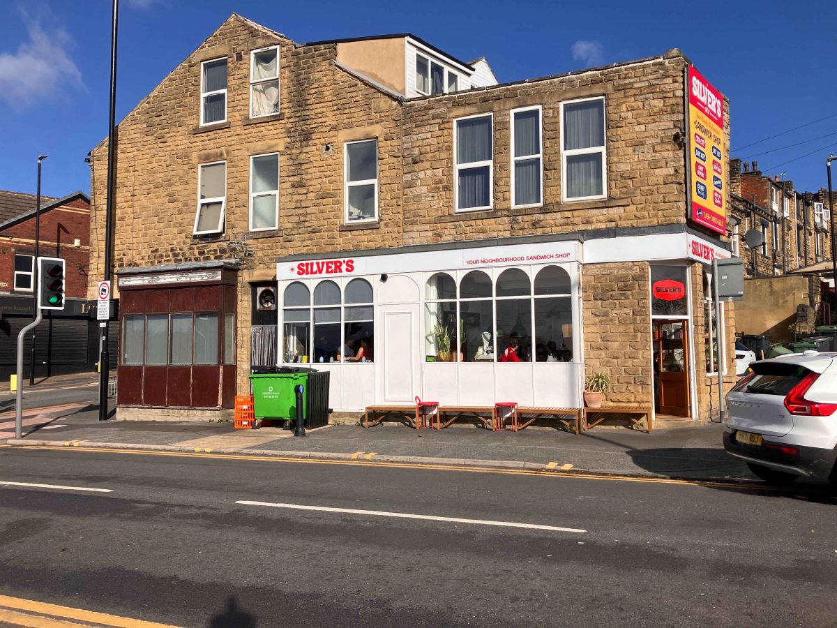 A street view of a stone building with a café on the ground floor named 'Silver's'. The café has a white façade with large arched windows and red chairs set up outside. Above the café is a bright red and yellow sign listing menu items like coffee, sandwiches, and breakfast. The building has multiple windows and a sloped roof, and there's a green waste bin and orange crates on the pavement. The sky is clear and blue.