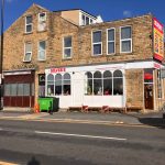 A street view of a stone building with a café on the ground floor named 'Silver's'. The café has a white façade with large arched windows and red chairs set up outside. Above the café is a bright red and yellow sign listing menu items like coffee, sandwiches, and breakfast. The building has multiple windows and a sloped roof, and there's a green waste bin and orange crates on the pavement. The sky is clear and blue.