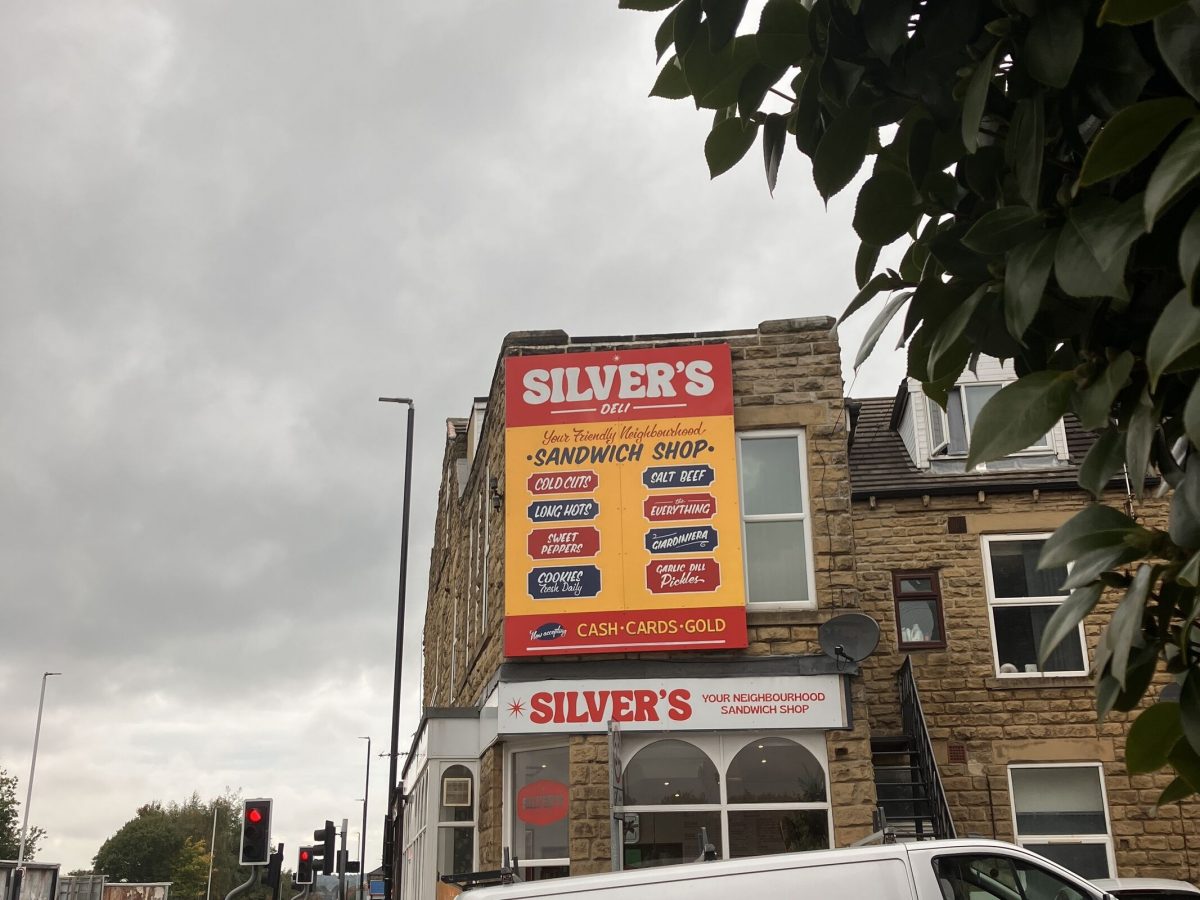 A photo of a stone building on a cloudy day featuring a large, colorful sign for "Silver's Deli" sandwich shop. The sign reads "Your Friendly Neighbourhood Sandwich Shop" and lists menu items such as cold cuts, long hots, salt beef, sweet peppers, everything, gorgonzola, garlic dill pickles, and cookies baked daily. The shop accepts cash, cards, and gold. A white van is parked in front, and a traffic light shows red in the background. Green leaves partially frame the right side of the image.