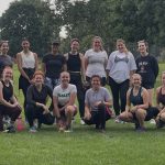 Thirteen women pose together outdoors on a grassy field, dressed in athletic wear. Some are kneeling in front while others stand behind, smiling and relaxed. Trees and a building are visible in the background. The group appears to have just finished a run.