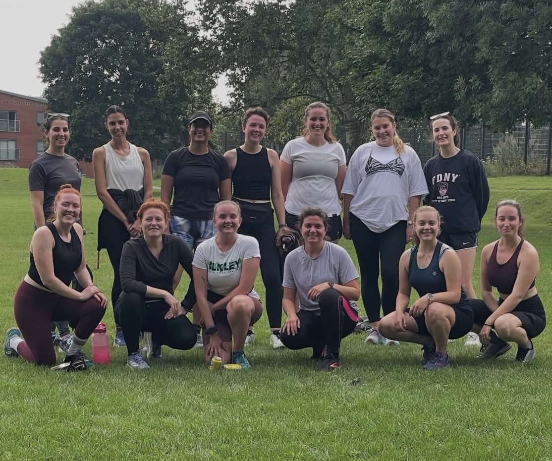 Thirteen women pose together outdoors on a grassy field, dressed in athletic wear. Some are kneeling in front while others stand behind, smiling and relaxed. Trees and a building are visible in the background. The group appears to have just finished a run.