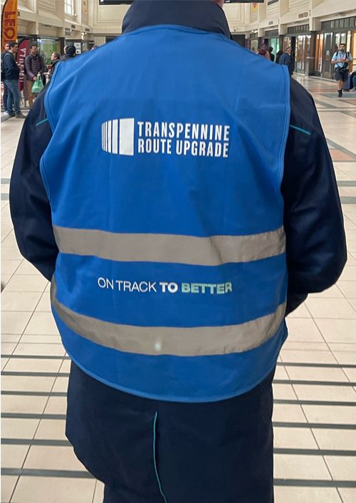 Person wearing a blue high-visibility vest with reflective stripes, printed with the logos and text ‘TRANSPENNINE ROUTE UPGRADE’ and the slogan ‘ON TRACK TO BETTER,’ standing inside a train station concourse.