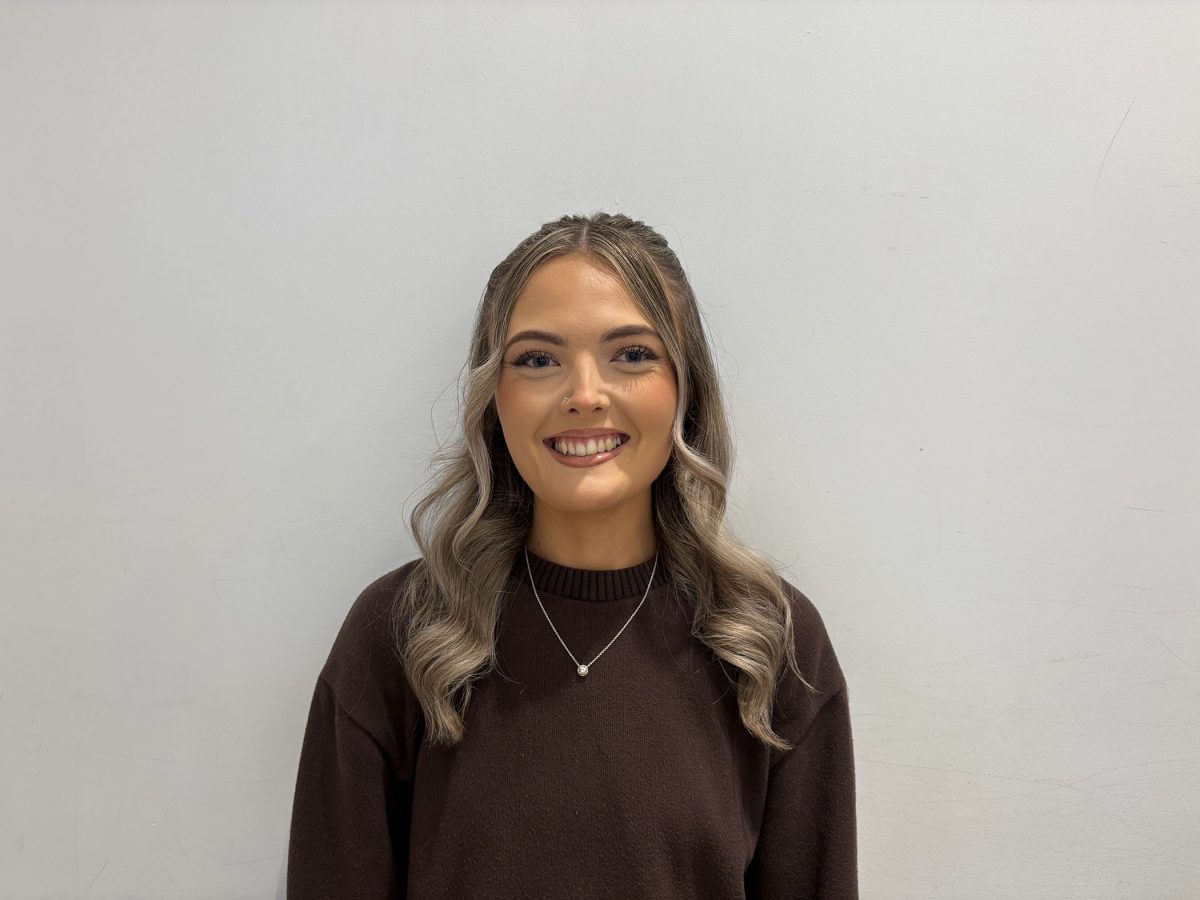 Young woman with long wavy hair, smiling warmly in front of a light gray background. She wears a dark brown sweater and a delicate necklace.