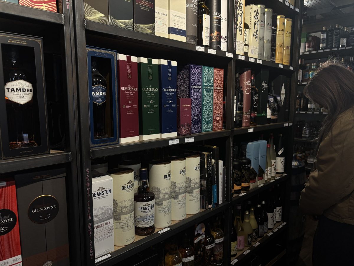 Shelves filled with neatly arranged bottles and boxes of whisky and liquor. A person wearing a brown jacket stands to the right.