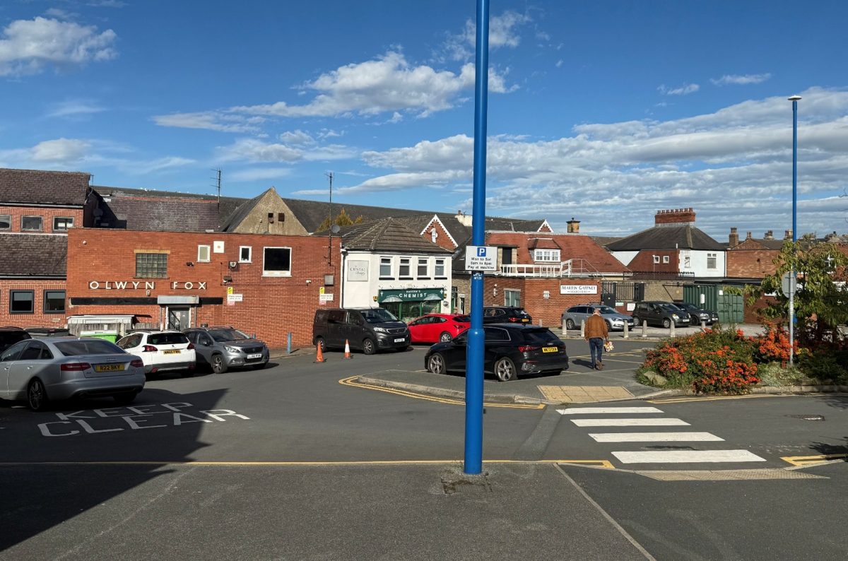 A small town car park on a sunny day with scattered clouds in the blue sky. Several cars are parked on the asphalt lot, including a silver Audi, a white hatchback, and a black SUV. The words “KEEP CLEAR” are painted on the ground. A tall blue pole with a parking sign in the center of the image states parking restrictions (Monday to Saturday, pay at machine). Behind the lot are a series of brick and white-painted buildings, including "OLWYN FOX" in bold letters on a red brick wall and “Naseem’s Chemist” visible on a white building. A pedestrian in a brown jacket walks away from the camera toward the far end of the car park. Flowering bushes with orange-red blossoms and a zebra crossing are in the foreground, and the background shows rooftops and chimneys of nearby buildings.