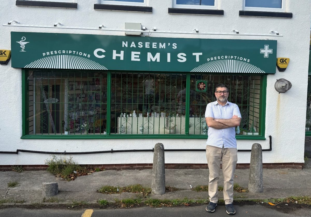 A man stands with his arms crossed outside a white building with green-framed windows and signage that reads "Naseem's Chemist – Prescriptions" in bold white letters on a green awning. The windows are barred with decorative metalwork, and inside the shop, various pharmacy items are visible. Above the entrance is a classic pharmacy symbol (a mortar and pestle), and a yellow security alarm box labeled "GK" is mounted on the wall. The man is wearing a short-sleeved checkered shirt, beige trousers, and dark trainers. The pavement in front has two stone bollards, patches of grass, and a yellow road marking.