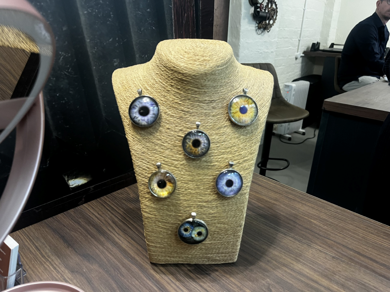 A display of six round pendants with colorful eye designs on a beige mannequin bust, set on a wooden table in a shop or studio, with chairs and a person at a desk in the background.