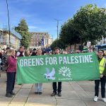 Four people holding up large green banner which reads "Greens for Palestine" with image of a white dove and the Green Party logo on it.