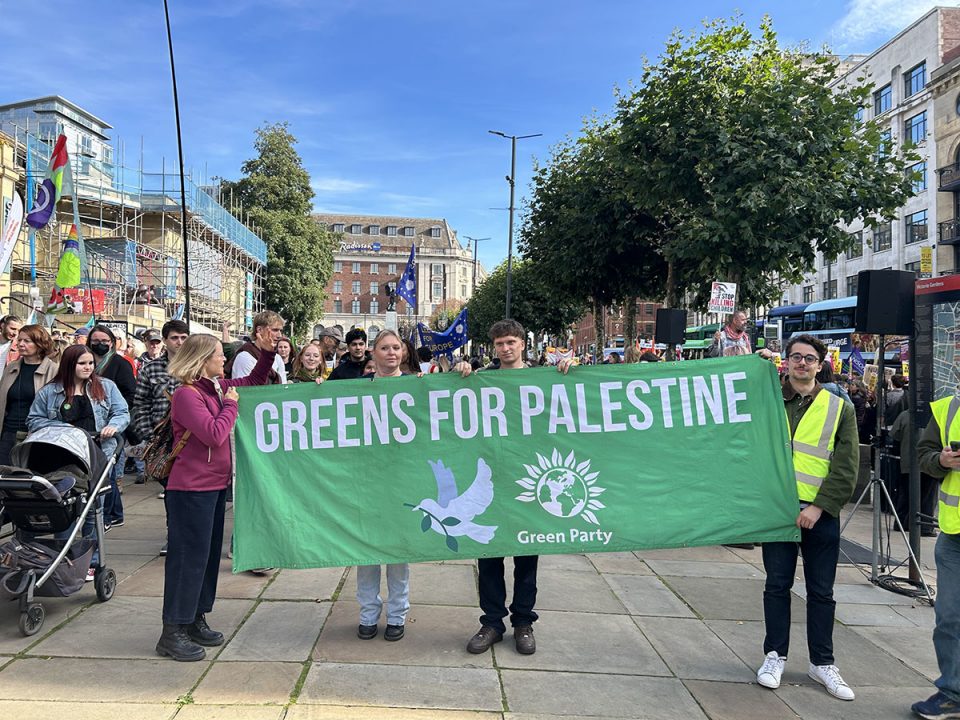 Four people holding up large green banner which reads "Greens for Palestine" with image of a white dove and the Green Party logo on it.