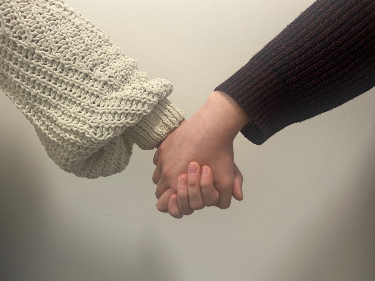 image of two white women holding hands against a white wall background. One woman is wearing a brown knit jumper and the other is wearing a cream knit jumper.