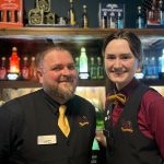 Benjamin Watkins and Emily Webb Two smiling staff members at The Hole in the Wand mini golf venue in Leeds, dressed in matching uniforms with black vests, yellow bow ties, and name tags, standing in front of a colourful potion-themed drinks display.