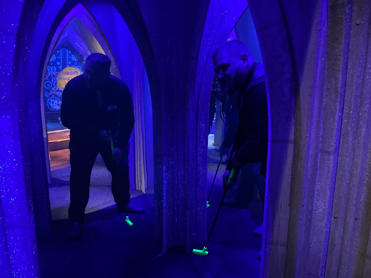 Benjamin Watkins playing mini golf in a dimly lit, fantasy-themed tunnel glowing with blue and purple lighting, holding a neon green putter. The surroundings resemble a magical castle with archways and glittering walls.