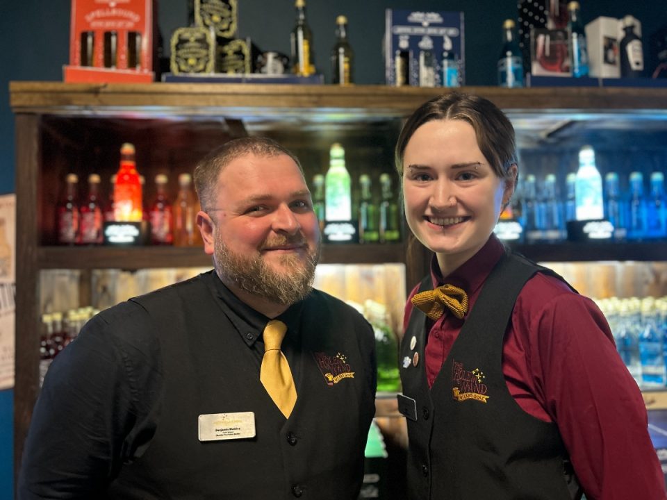 Benjamin Watkins and Emily Webb Two smiling staff members at The Hole in the Wand mini golf venue in Leeds, dressed in matching uniforms with black vests, yellow bow ties, and name tags, standing in front of a colourful potion-themed drinks display.