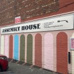 image of building with pink painted walls and painted multi-colour arches. A white sign hangs across the wall saying Assembly House.