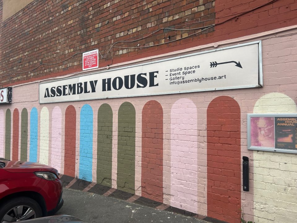 image of building with pink painted walls and painted multi-colour arches. A white sign hangs across the wall saying Assembly House.