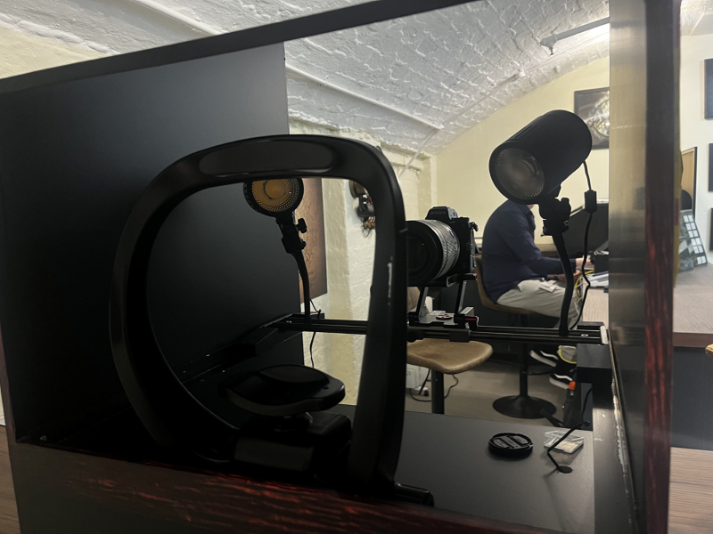  A photography setup in a room with arched ceilings, featuring a ring light, a camera on a tripod, and another cylindrical light source, with a person working at a desk in the background.