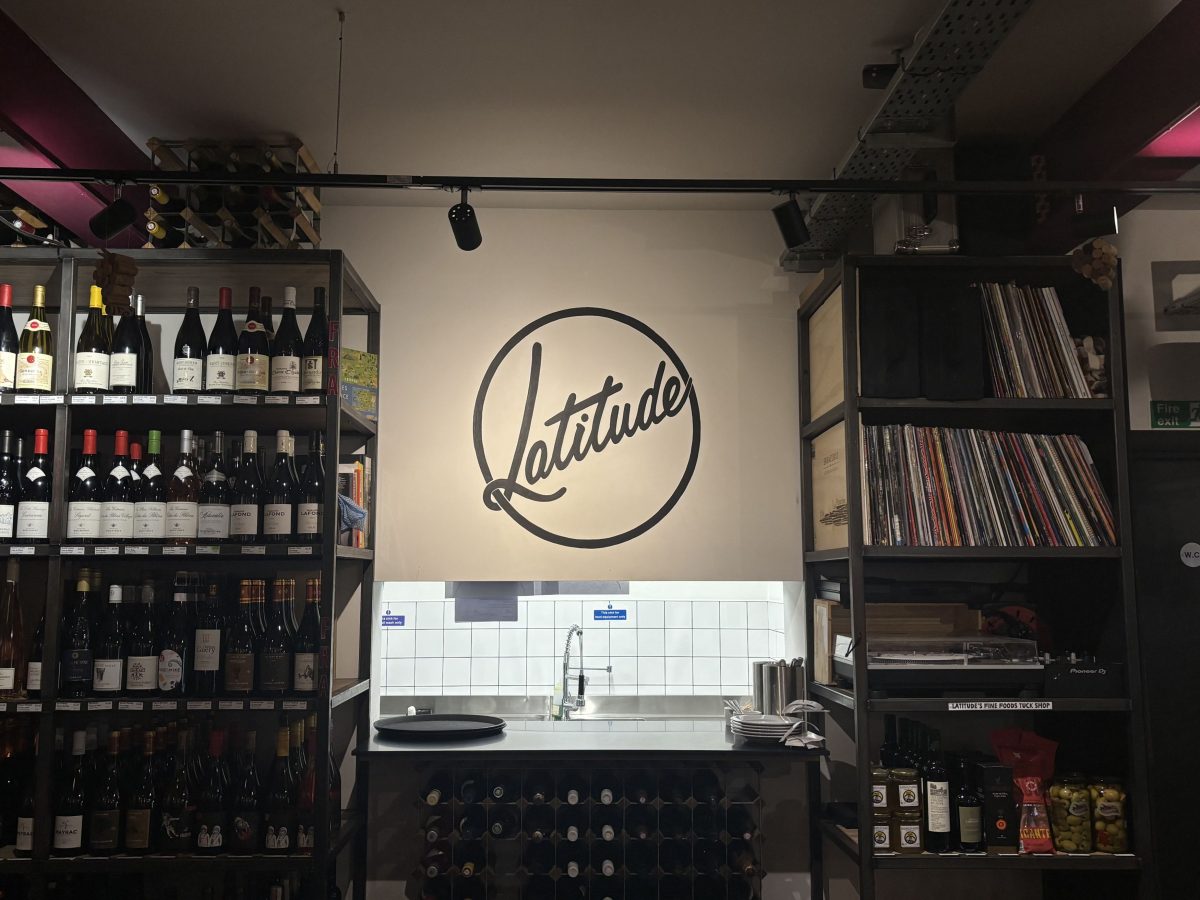 The image shows the interior of a wine and spirits shop named “Latitude.” At the center is a wall with a circular logo that reads “Latitude” in cursive script. Below the logo is a small sink area with white tiled walls, a stainless steel countertop, and some kitchen utensils and plates. On either side of the wall are shelves: the left side is filled with various bottles of wine, and the right side holds a large collection of vinyl records along with a turntable and other items like jars of olives and packaged goods. There is a wine rack below the countertop holding multiple wine bottles.
