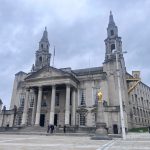 Leeds Civic Hall, a grand neoclassical building with two tall clock towers topped by golden owls, stands prominently in a public square. The façade features white stone columns and ornate carvings. In the foreground, a golden statue and a decorative clock add visual interest, while a few pedestrians walk across the square under an overcast sky.