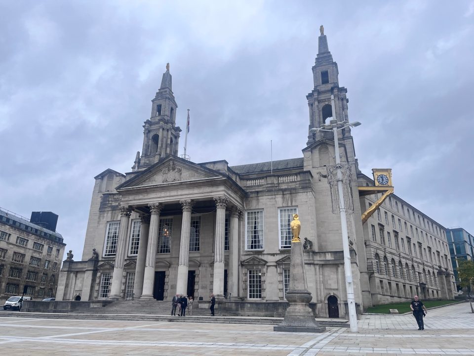 Leeds Civic Hall, a grand neoclassical building with two tall clock towers topped by golden owls, stands prominently in a public square. The façade features white stone columns and ornate carvings. In the foreground, a golden statue and a decorative clock add visual interest, while a few pedestrians walk across the square under an overcast sky.