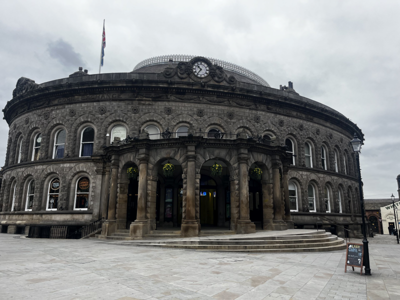  A historic round stone building with a domed roof, arched windows, and a clock above the entrance, with a flag flying on top and an overcast sky in the background.