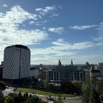 Cityscape view of modern buildings under a bright blue sky with scattered clouds. Green trees and a busy intersection add urban vibrancy.