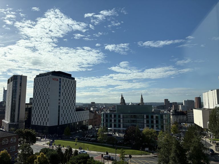 Cityscape view of modern buildings under a bright blue sky with scattered clouds. Green trees and a busy intersection add urban vibrancy.