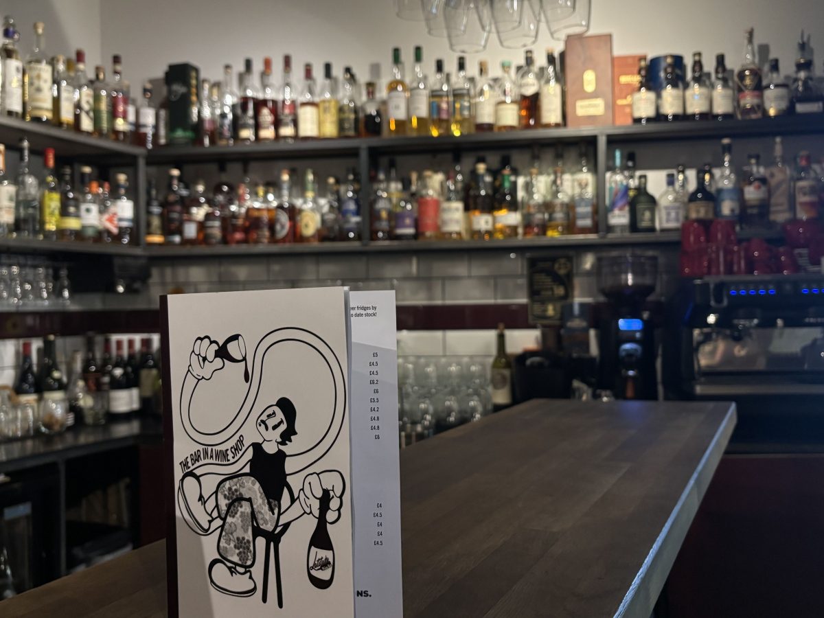 A cozy bar area inside a wine and liquor shop, with shelves lined with bottles. A wooden bar counter sits in the foreground, displaying a menu with an illustration of a person drinking wine and the text "The Bar in a Wine Shop."