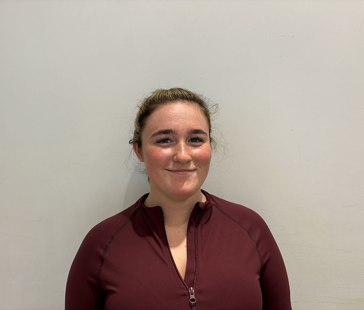 Young person with light brown hair tied back, smiling slightly, wearing a maroon zip-up top, standing against a plain off-white background.